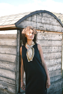 Woman wearing block print Savanna bandana tied around neck against a black sleeveless dress. Woman leaning against a wooden building.