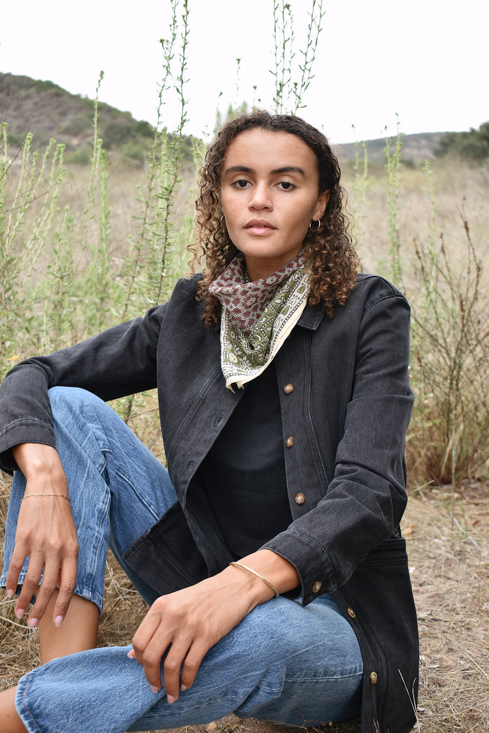Model wearing Janis bandana, tied in the back around neck, worn with black denim jacket and blue jeans. Model is sitting outside in a field.