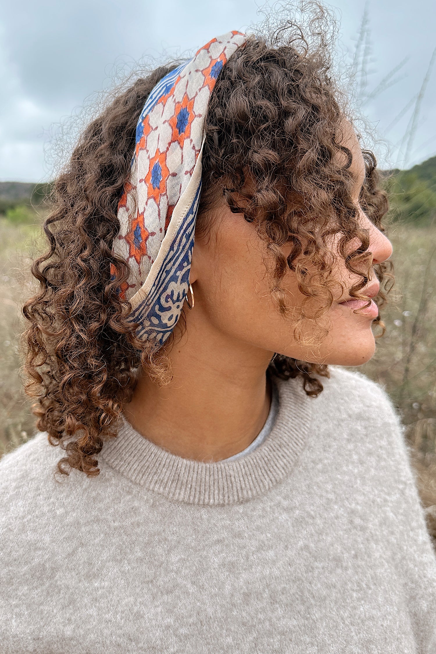 Woman wearing a tan sweater, standing in a field, with the block print Jane cotton bandana tied around her hair as a headband.