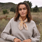 Model wearing block printed yellow Suki bandana around the neck, paired with tan sweater, blue jeans. Model standing outside around trees, against a fence.