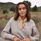 Model wearing block printed yellow Suki bandana around the neck, paired with tan sweater, blue jeans. Model standing outside around trees, against a fence.