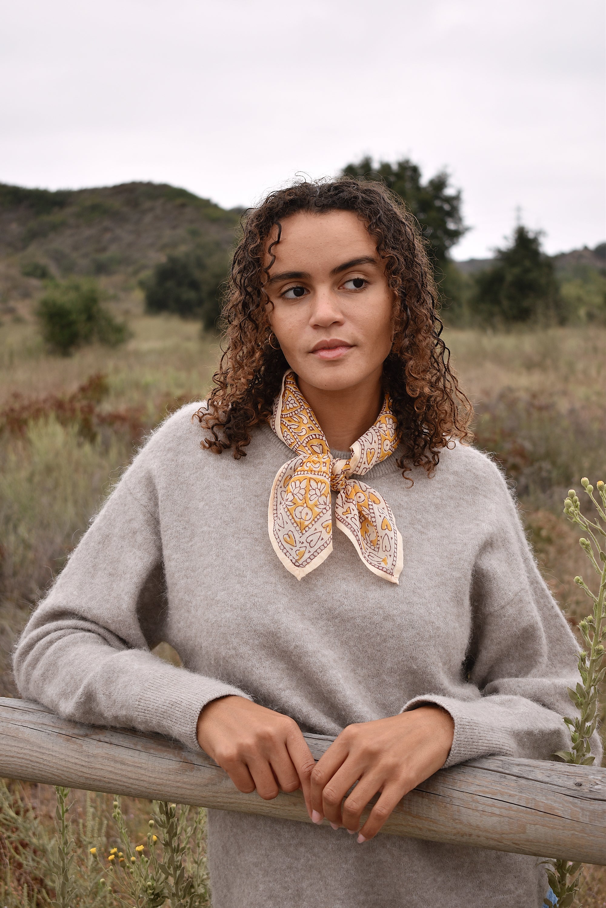 Model wearing block printed yellow Suki bandana around the neck, paired with tan sweater, blue jeans. Model standing outside around trees, against a fence.