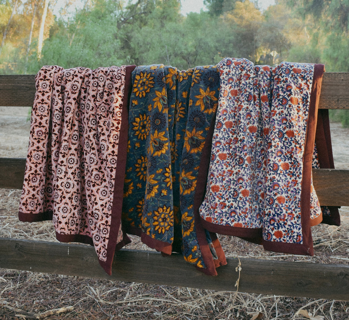 Three patterned blankets draped over a rustic wooden fence outdoors, with trees and dry grass visible in the background.