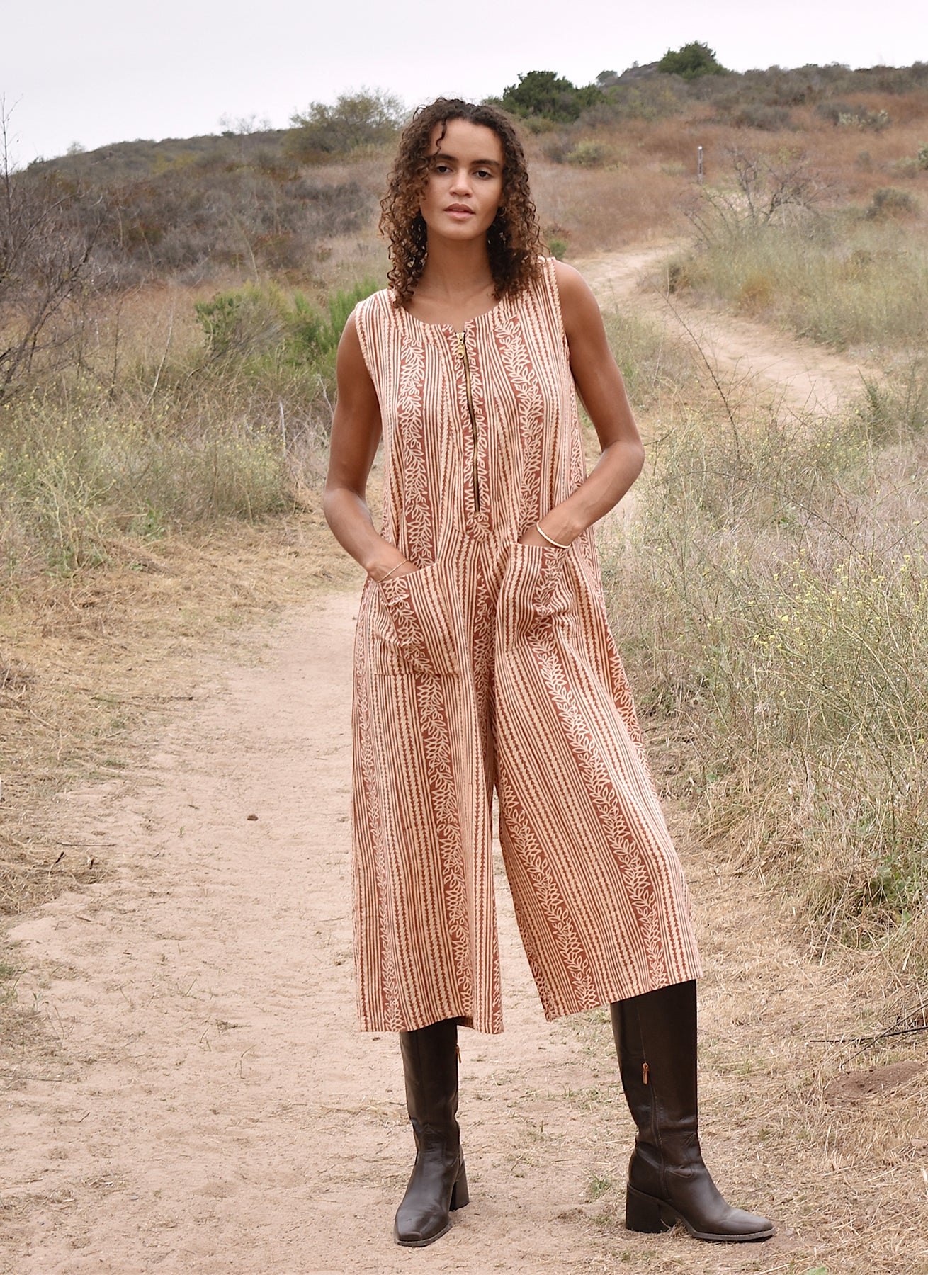 Model wearing block printed sleeveless Taylor jumpsuit with brown boots outside, standing on a hill