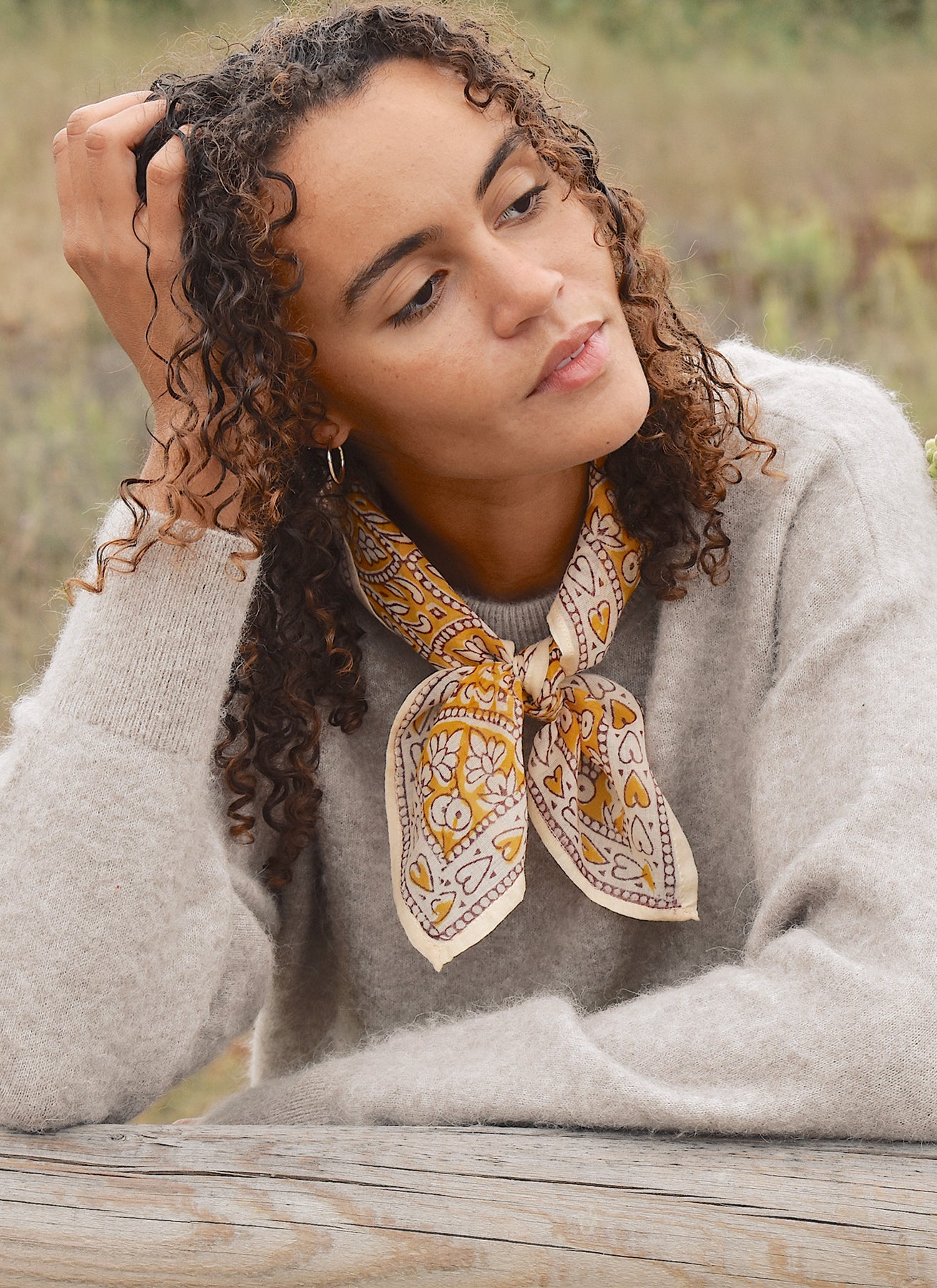 Woman in tan sweater wearing block print yellow and white Suki Bandana around her neck. Woman leaning against a wooden fence with field/grass in the background.