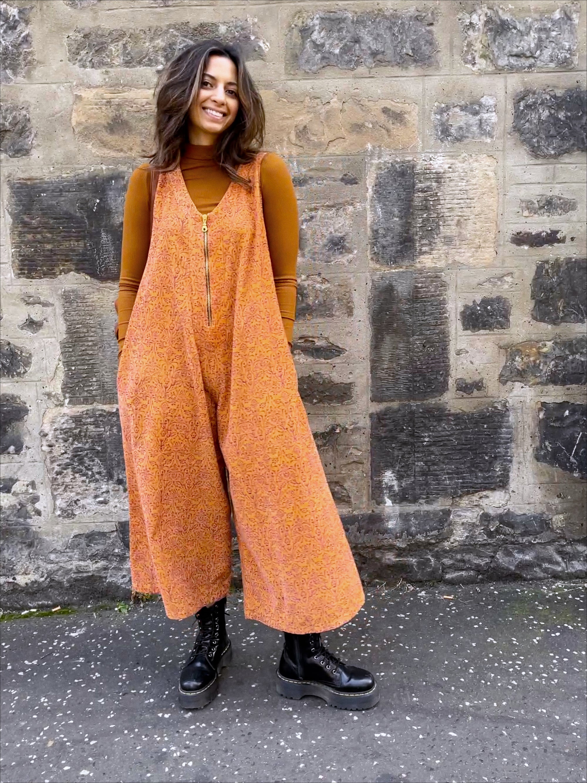 Woman wearing Stevie Jumpsuit, layered with a turtleneck and paired with boots, in front of building