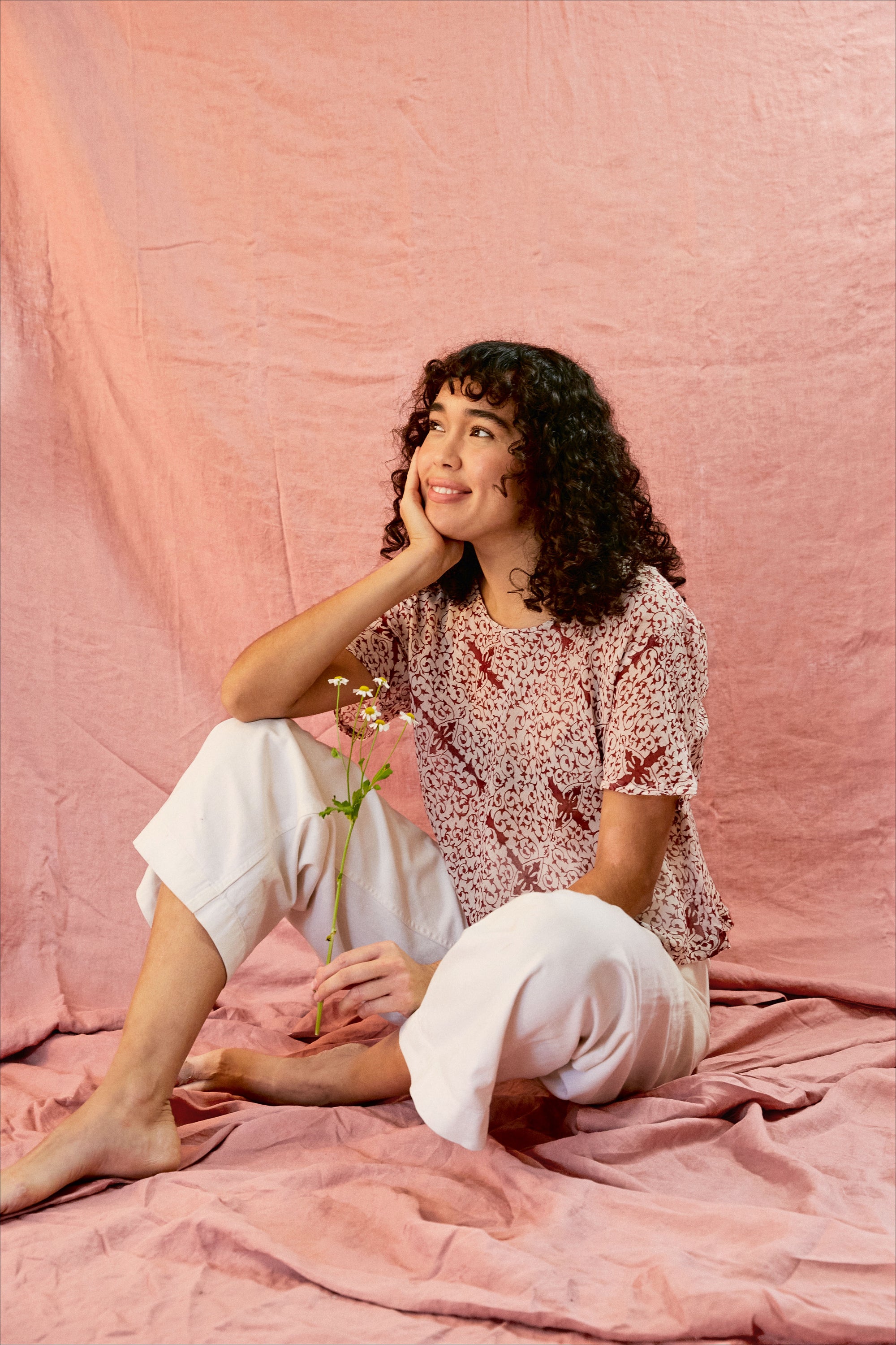 Model wearing the Alicia Top with white pants, against a pink backdrop. Model is holding a flower and sitting down.