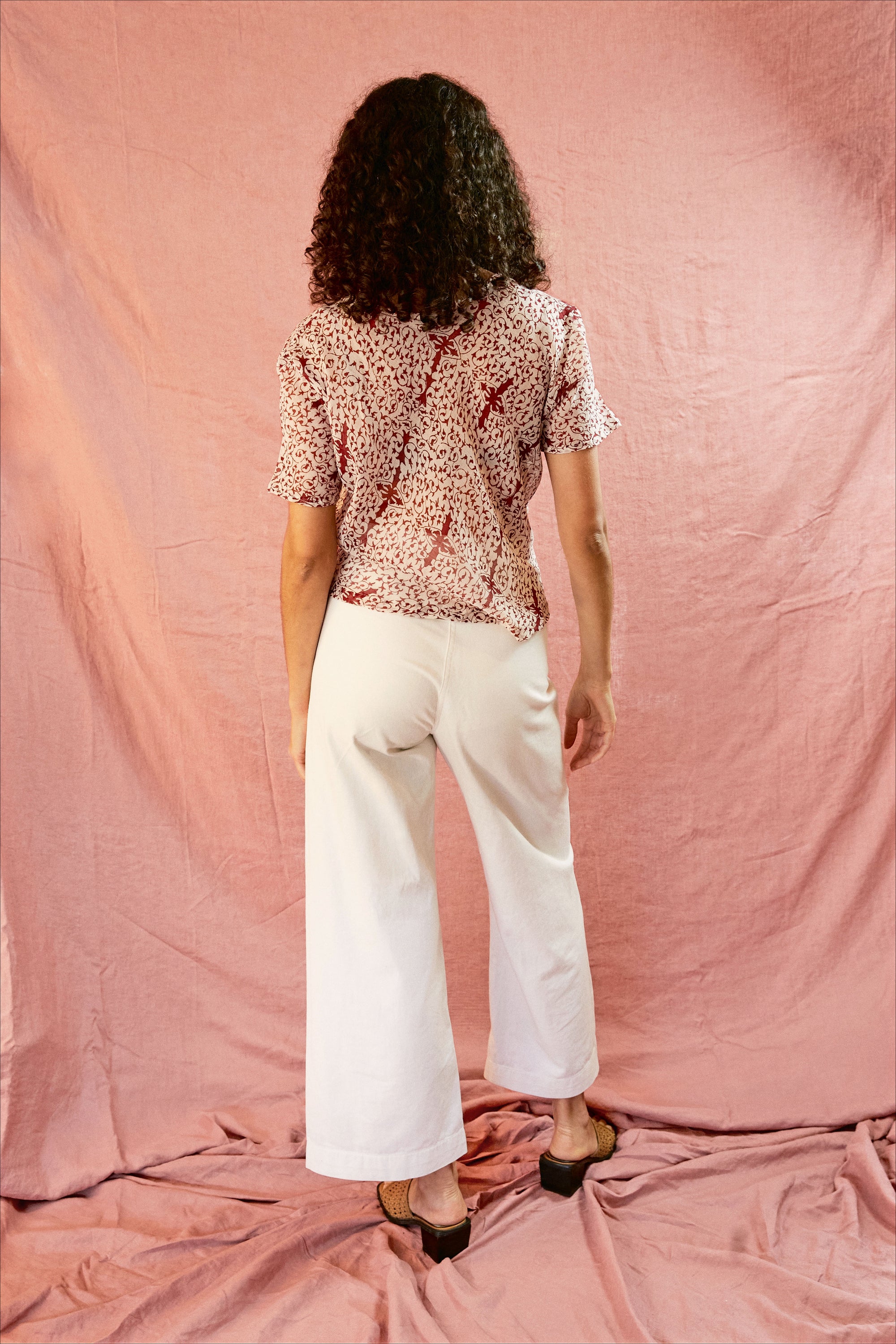 Model wearing the Alicia Top with white pants, against a pink backdrop. Backside of model.