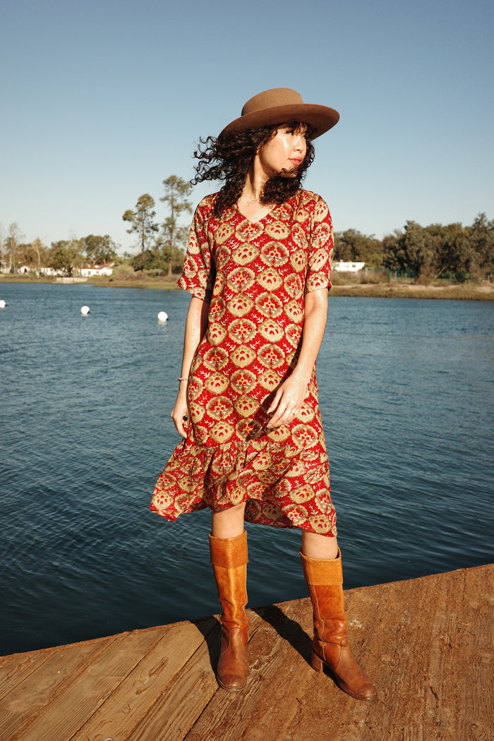 Model wearing the Sienna Ruffle Dress in the Sintra print with boots and a hat near a body of water
