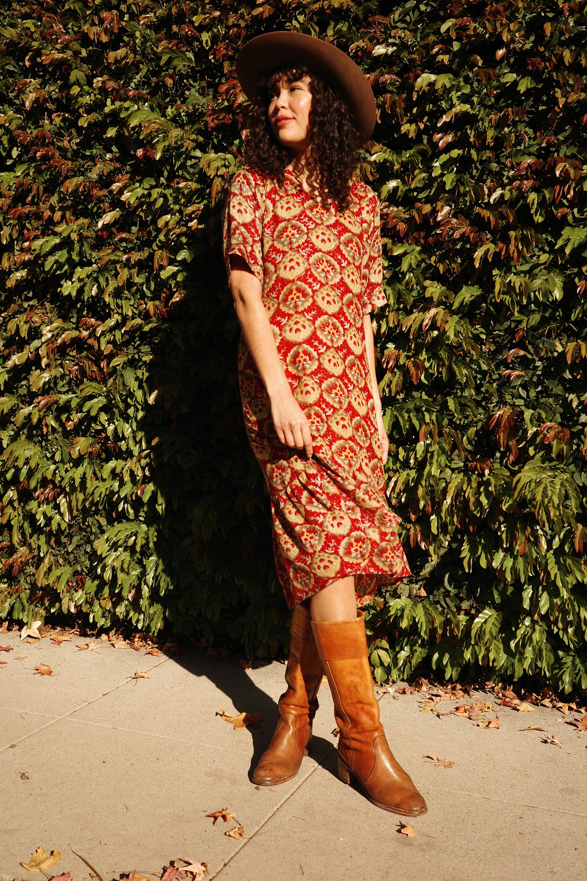 Model wearing the Sienna Ruffle Dress in the Sintra print with boots and a hat in front of a tall bush