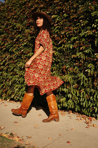 Model walking in the Sienna Ruffle Dress in the Sintra print with boots and a hat in front of a tall bush