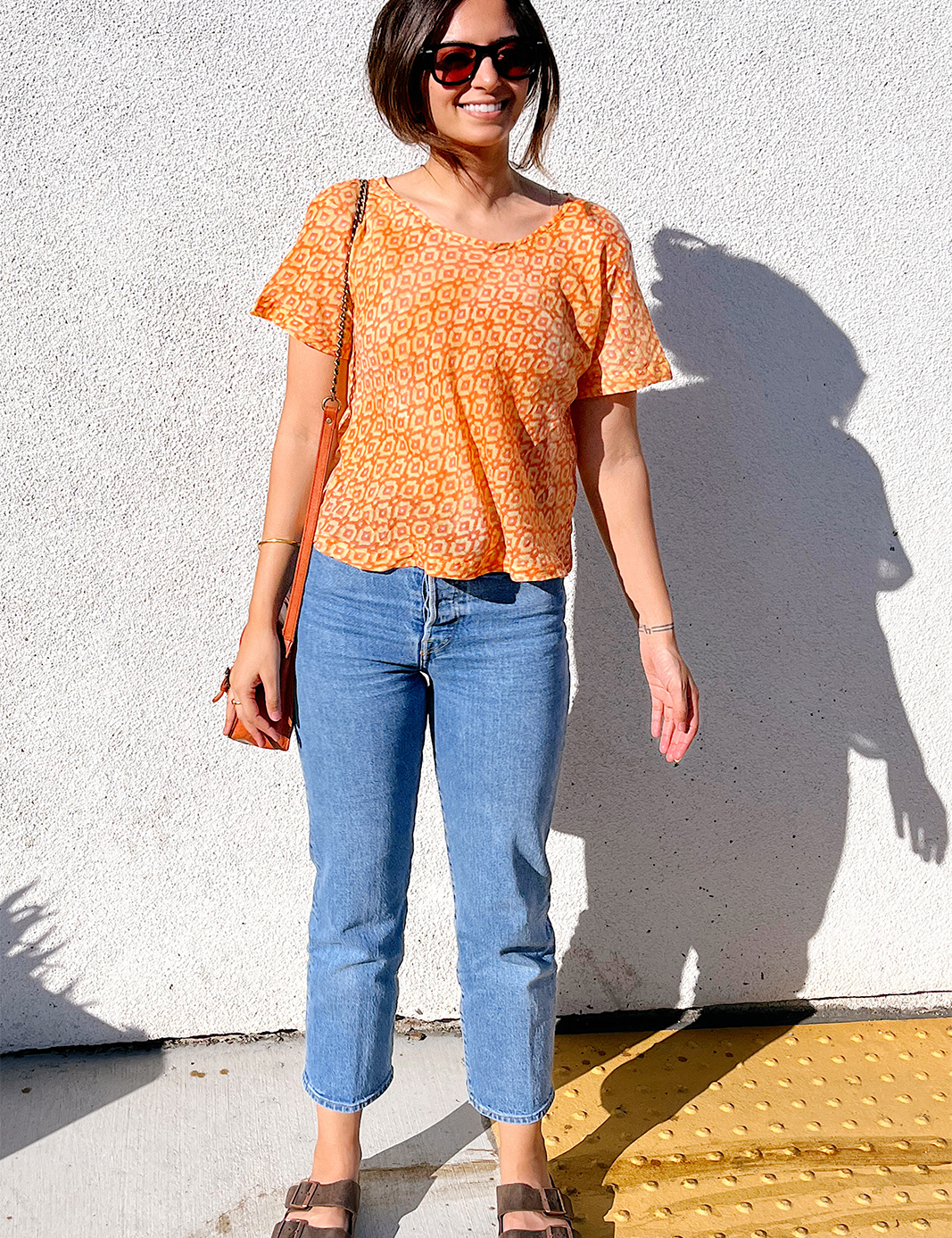 Woman is wearing the Sophie Crop Top with a cropped pair of jeans, sandals and a purse. Against a white wall.